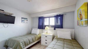 Une chambre avec deux lits simples, une table de chevet avec une lampe, un téléviseur à écran plat fixé au mur et une fenêtre avec des rideaux.