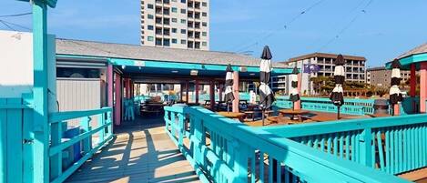 A wooden pier with turquoise railings, outdoor seating, and a view of a tall building in the background.