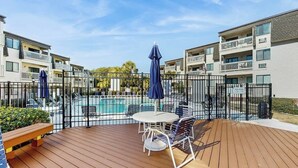 A wooden deck with outdoor furniture, a swimming pool, and apartment buildings in the background. - Ocean Forest Villas A-216 (Myrtle Beach)