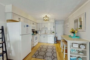 A compact kitchen with white cabinets, a refrigerator, a microwave, and a sink. There's a patterned rug on the floor and a small table with a vase of flowers. - Ocean Forest Villas A-216 (Myrtle Beach)