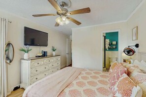 Ein Schlafzimmer mit Bett, Deckenventilator, Kommode, Fernseher und einer Tür zu einem weiteren Raum.
