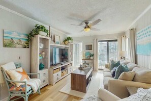 Ein Wohnzimmer mit Fernseher, Sofa und Sessel. Es gibt einen Deckenventilator, ein Fenster mit Vorhängen und eine Pflanze auf einem Regal.
