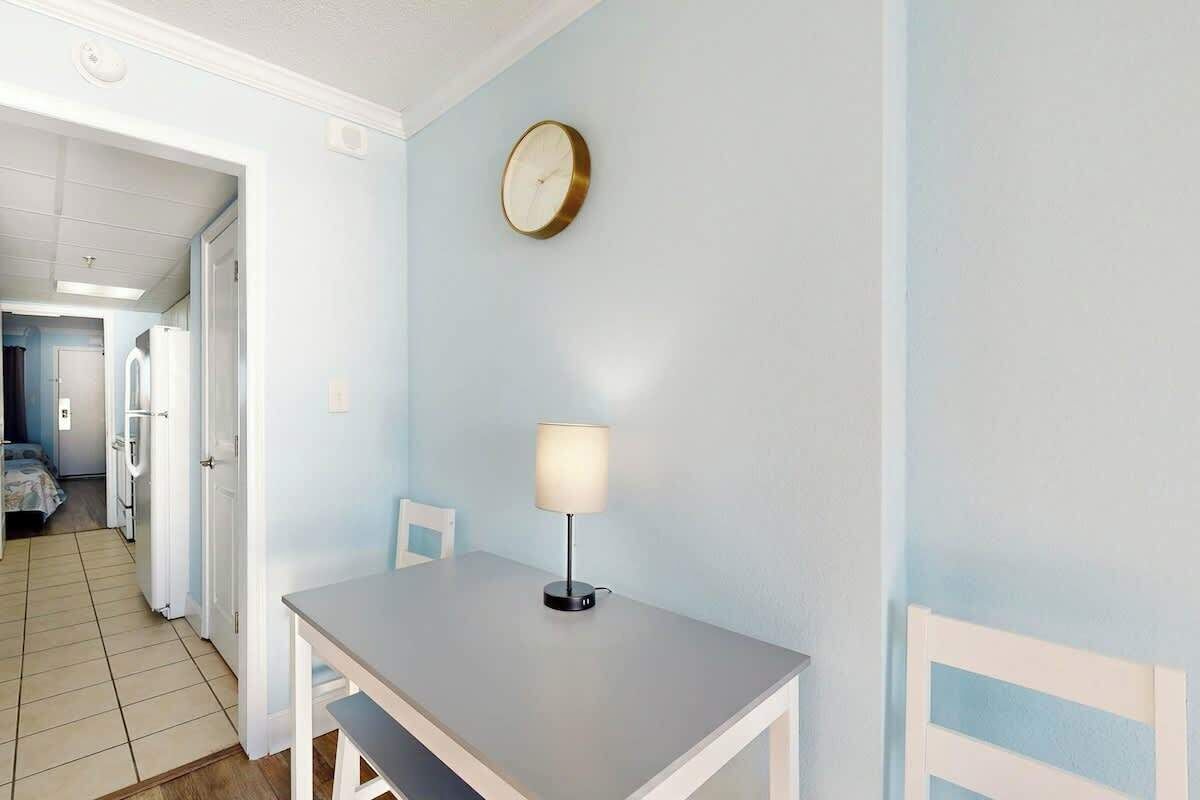 A room with a table, a lamp, a clock, and a door leading to another room.