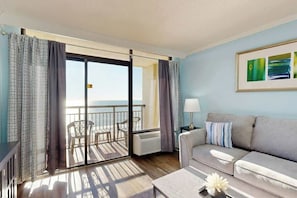 A room with a sofa, a table with a vase, a painting, and a view of the sea through a sliding door. - Here's Your 5 O'Clock Somewhere (Myrtle Beach)