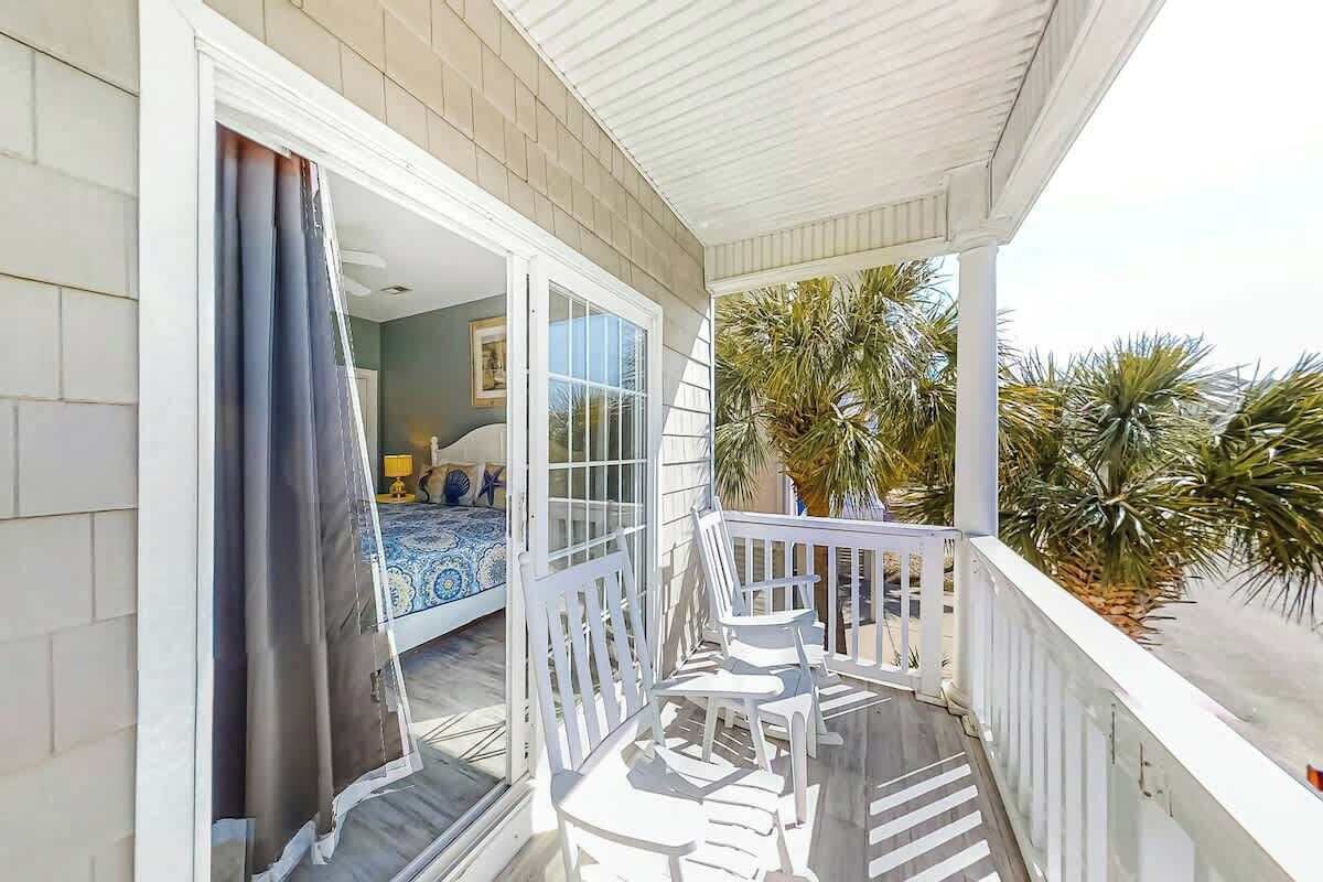A balcony with a white railing, a bed with a blue and white patterned bedspread, and a view of palm trees.