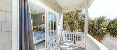 A balcony with a white railing, a bed with a blue and white patterned bedspread, and a view of palm trees.