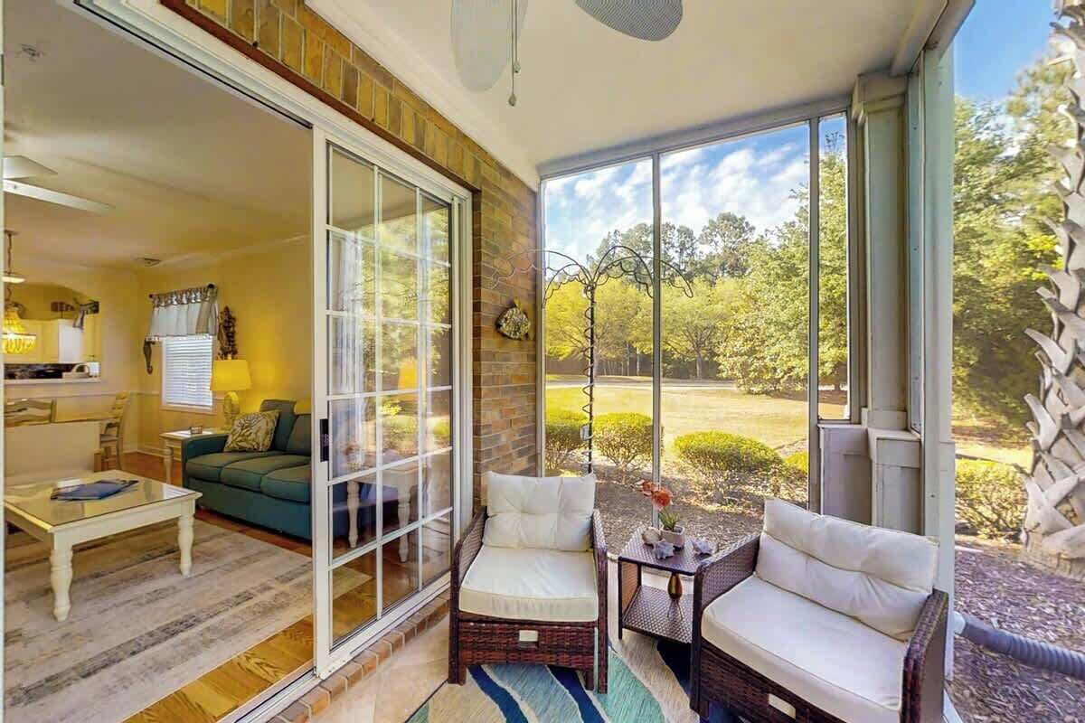 A sunlit room with a sliding glass door, a green sofa, a white armchair, and a small table with a vase.