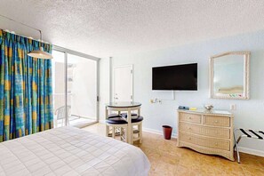 Ein Hotelzimmer mit einem Bett, einem kleinen Tisch mit Stühlen, einem Fernseher, einem Schminktisch und einer Glasschiebetür, die auf einen Balkon führt.