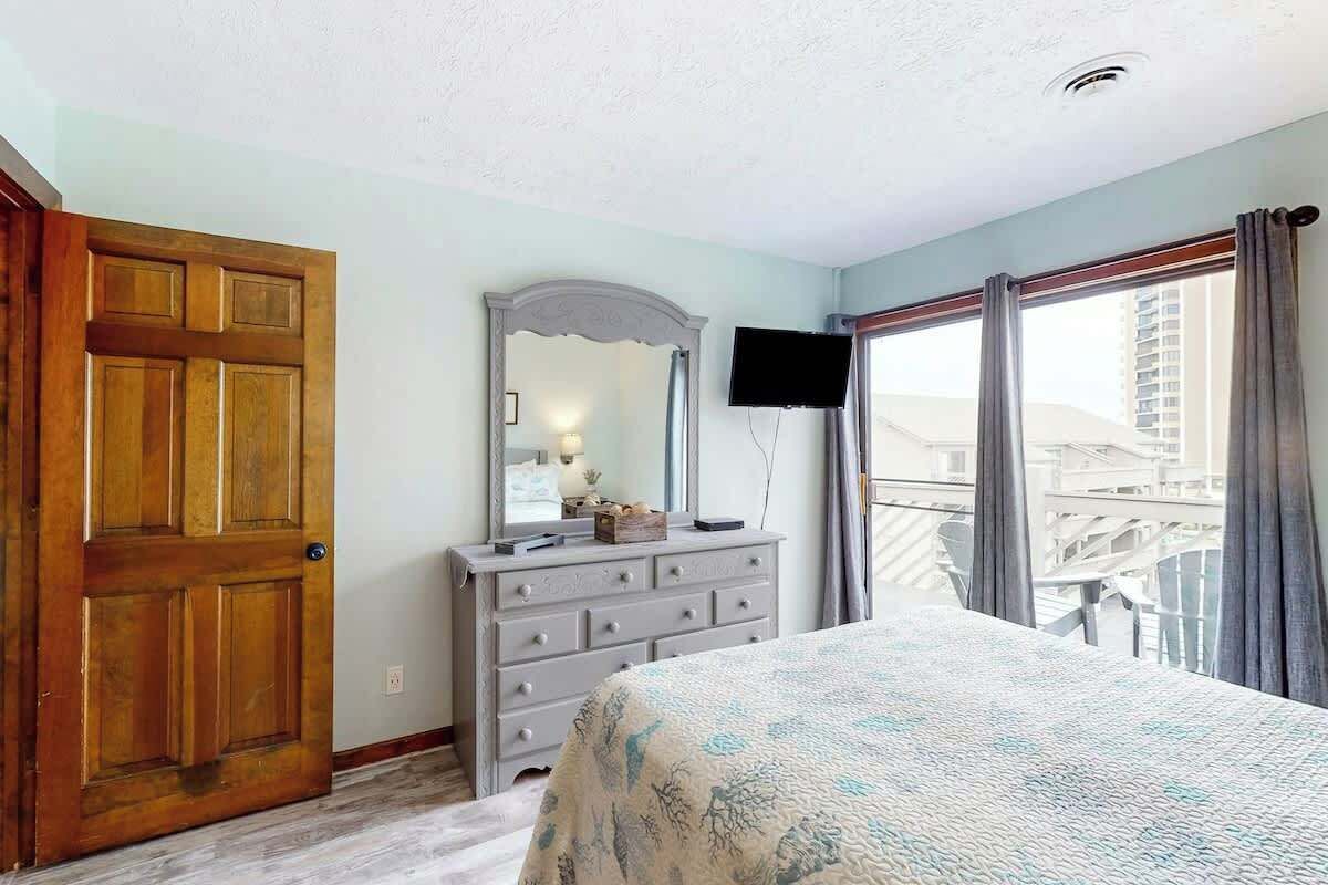 A bedroom with a wooden door, a dresser, a bed with a patterned bedspread, a television, and a window with curtains.