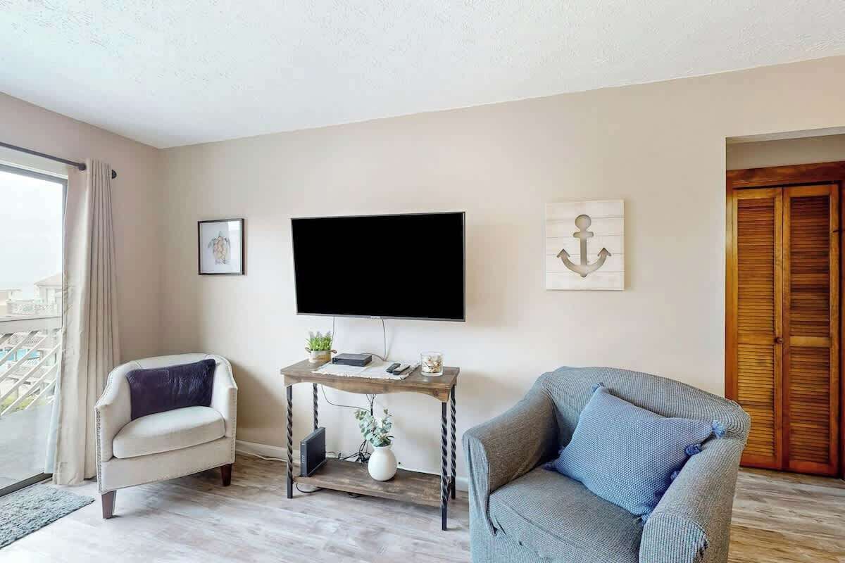 A living room with a flat-screen TV mounted on the wall, a small wooden side table with a plant, a grey sofa, and a white armchair.