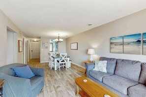 A living room with a grey sofa, a wooden coffee table, and a dining area with a white table and chairs. - Shipwatch Pointe I E-308 (Myrtle Beach)