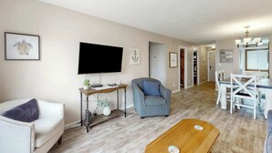 A living room with a flat-screen TV, a sofa, a chair, a coffee table, and a dining area with a table and chairs.