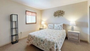 A bedroom with a bed, two bedside tables, a nightstand, a wall decoration, a window with a valance, and a tall black shelf. - Water's Edge Resort 1501 (Murrells Inlet)