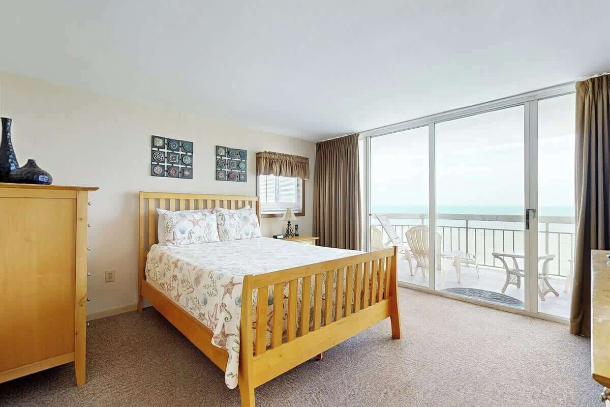 A hotel room with a bed, a desk, a chair, a window with a view of the sea, and a balcony with outdoor furniture.