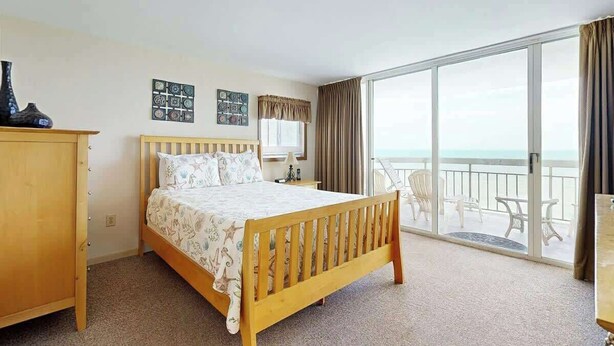 A hotel room with a bed, a desk, a chair, a window with a view of the sea, and a balcony with outdoor furniture. - Water's Edge Resort 1501 (Murrells Inlet)