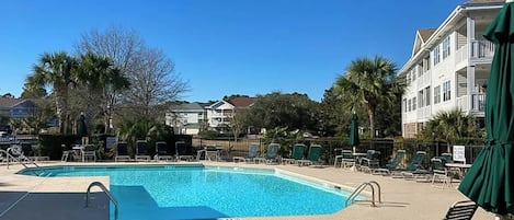 Ein Pool mit Liegestühlen und Sonnenschirmen, umgeben von Gebäuden und Palmen.