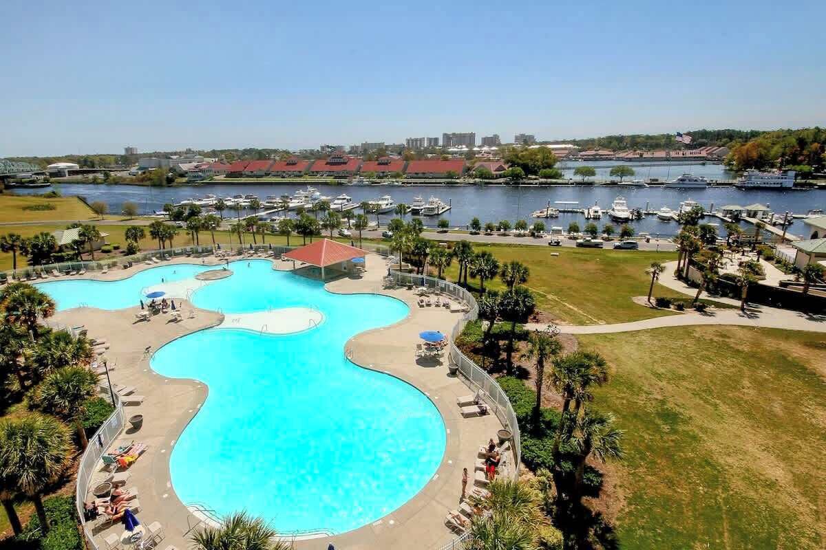 A large, curved swimming pool with lounge chairs and umbrellas, surrounded by a grassy area and palm trees.