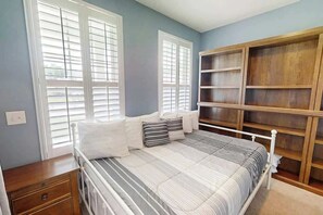 A bedroom with a bed, bedside tables, a built-in wooden shelving unit, and windows with blinds. - Turtle Cove Beach Retreat (Myrtle Beach)