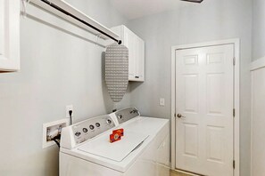 A laundry room with a front-loading washer and dryer, a wall-mounted ironing board, and a white door. - Turtle Cove Beach Retreat (Myrtle Beach)