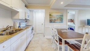 A modern kitchen with white cabinets, a granite countertop, and a dining area with a wooden table and white chairs. - Summerhouse at Litchfield (Pawleys Island)