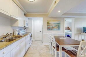 A modern kitchen with white cabinets, a granite countertop, and a dining area with a wooden table and white chairs. - Summerhouse at Litchfield (Pawleys Island)