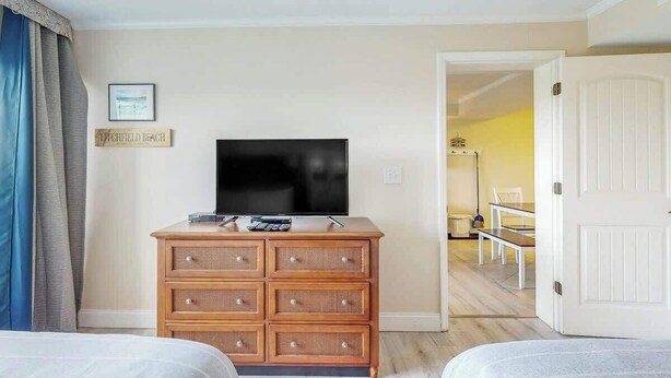 A hotel room with a television on a dresser, a bed, and a bench in the adjacent room. - Summerhouse at Litchfield (Pawleys Island)