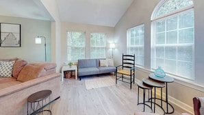 A living room with a sofa, a chair, a small table, and a lamp. There are windows with blinds and a chandelier. - Seascape Beach Retreat (Murrells Inlet)