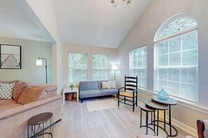Ein Wohnzimmer mit einer Couch, einem Sessel, einem kleinen Tisch und einer Lampe. Es gibt Fenster mit Jalousien und eine Kristallleuchter.