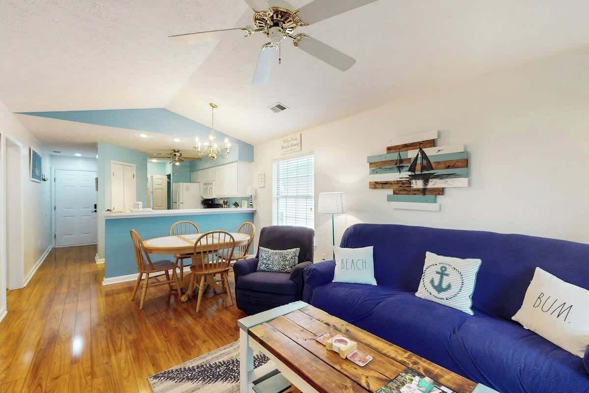 Un soggiorno con un divano blu, un tavolo da pranzo con sedie, una ventola a soffitto e una cucina sullo sfondo.
