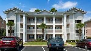 A multi-story residential building with a central courtyard, balconies, and a parking lot with cars. - Anchors Away Unit 7G (Myrtle Beach)
