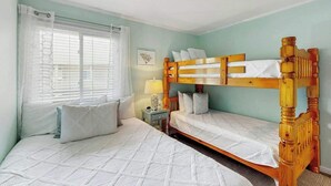 Un dormitorio con dos camas, una ventana con persianas, una mesita de noche con lámpara y un cuadro en la pared.
