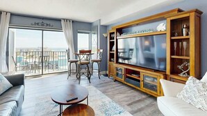 A living room with a large flat-screen TV, a wooden entertainment unit, a balcony with outdoor seating, and a view of the city. - Sunrise at the Beach (Myrtle Beach)