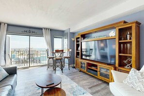 A living room with a large flat-screen TV, a wooden entertainment unit, a balcony with outdoor seating, and a view of the city. - Sunrise at the Beach (Myrtle Beach)