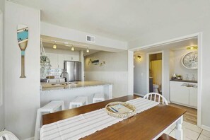A dining area with a wooden table, white chairs, and a kitchen in the background. - Palmetto Dunes Unit 303 (North Myrtle Beach)