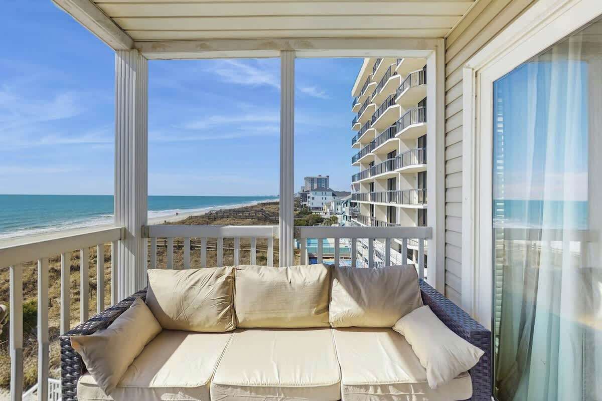 A balcony with a cushioned seating area overlooking a beach and a hotel building.