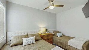 Ein Schlafzimmer mit zwei Betten, einem Deckenventilator, einem Nachttisch mit Lampe und einem an der Wand montierten Fernseher.