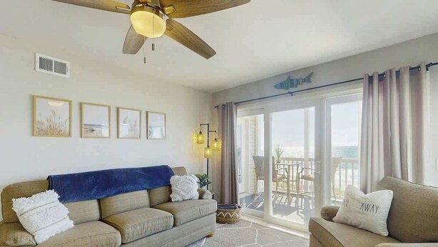 Ein Wohnzimmer mit Sofa, Sessel, Deckenventilator und Blick durch eine Glastür ins Freie.