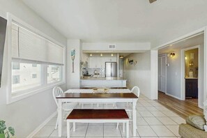 A dining area with a table and chairs, a kitchen in the background, and a view through a window. - Palmetto Dunes Unit 303 (North Myrtle Beach)