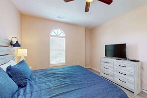 Ein Schlafzimmer mit einem Bett, einem Fernseher auf einer Kommode, einem Deckenventilator und einem Fenster mit Jalousien.