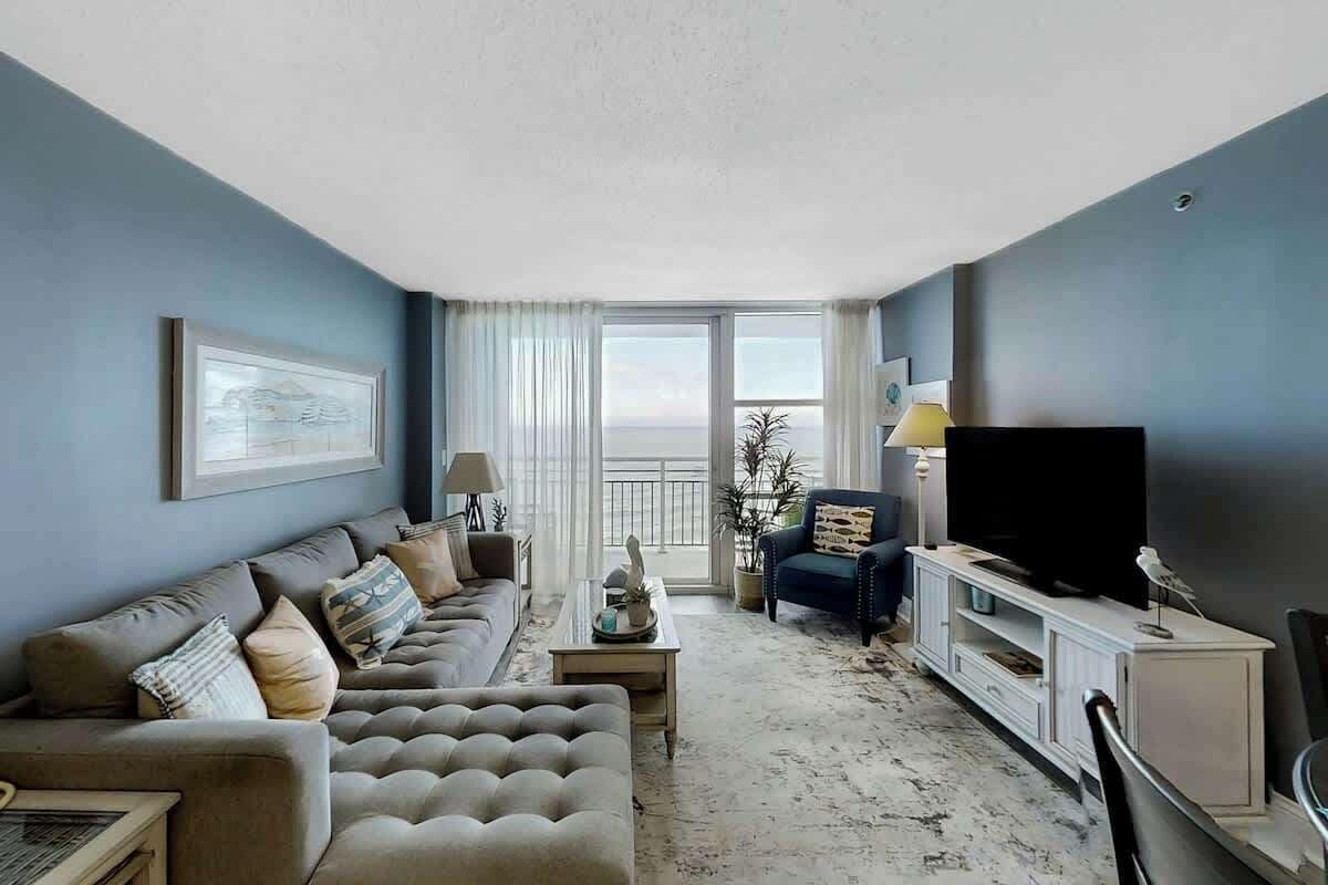 Una sala de estar con sofá, mesa de centro y televisor. Se aprecia una ventana grande con vistas.