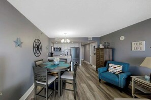 A modern living room with a dining area, kitchen in the background, and a blue sofa. - This is It 2 BR Condo (North Myrtle Beach)