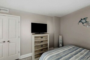 Quarto com uma cama, uma televisão sobre uma cômoda de madeira, um letreiro de boas-vindas e uma decoração na parede.