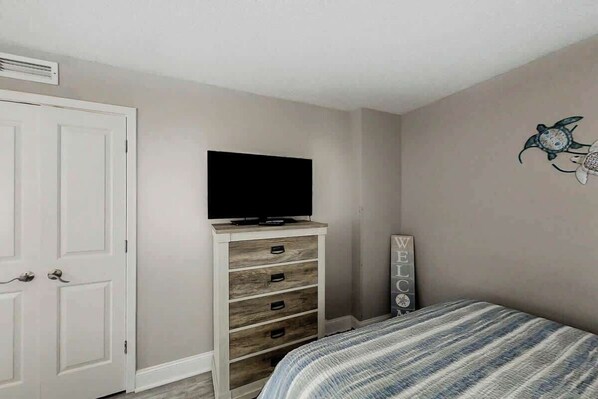A bedroom with a bed, a television on a wooden dresser, a welcome sign, and a wall decoration. - This is It 2 BR Condo (North Myrtle Beach)