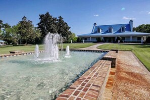 A clear outdoor pool with a fountain, surrounded by a brick walkway, and a large blue building in the background.