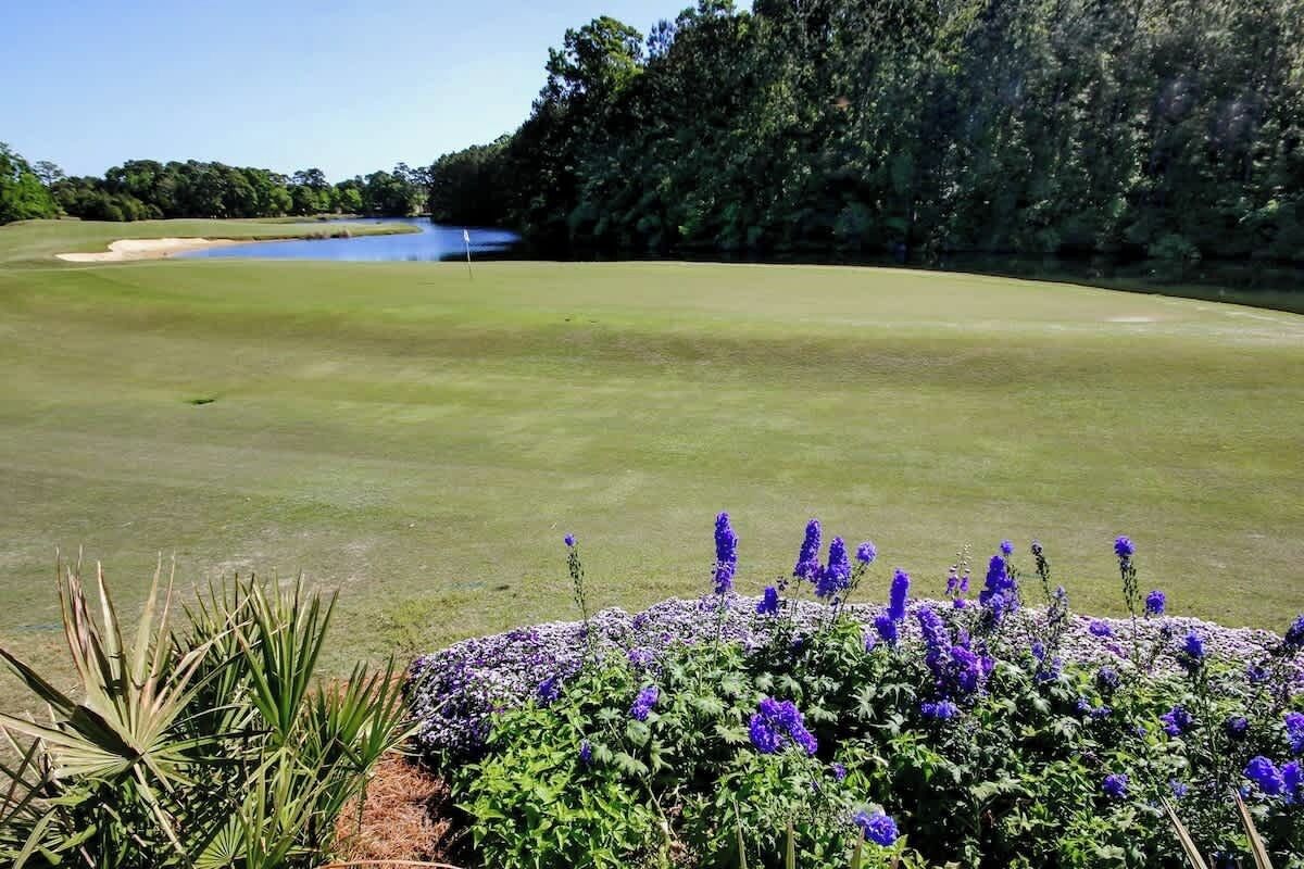 Een golfbaan met een waterhindernis, omringd door dichte bomen en paarse bloemen op de voorgrond.
