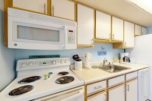 A kitchen with a white stove, microwave, and sink. There are wooden cabinets and a light blue wall. - Bay Watch Resort 1103 (North Myrtle Beach)