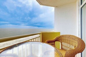 A balcony with a wicker chair and a glass table, overlooking a beach. - Bay Watch Resort 1103 (North Myrtle Beach)