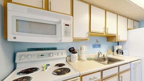 A kitchen with a white stove, microwave, and sink, wooden cabinets, and a coffee maker. - Bay Watch Resort 1103 (North Myrtle Beach)