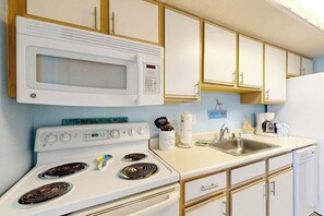 A kitchen with a white stove, microwave, and sink, wooden cabinets, and a coffee maker. - Bay Watch Resort 1103 (North Myrtle Beach)