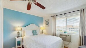 A bedroom with a bed, two bedside lamps, a ceiling fan, a window with curtains, and a view of the outside. - Coastal Tranquility Retreat (North Myrtle Beach)